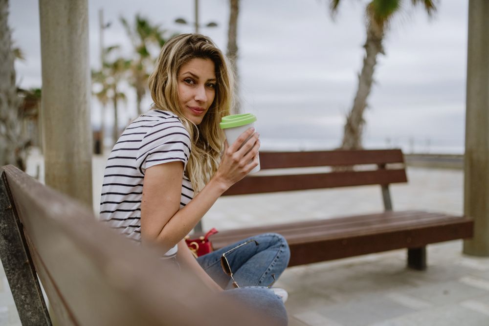 Happy woman with disposable cup of coffee sitting on street bench