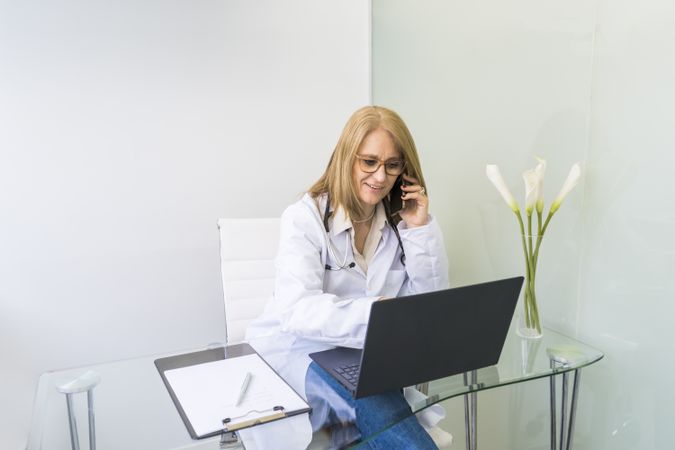 Smiling female doctor talking on the phone to a patient while looking at her medical records on laptop in medical office.