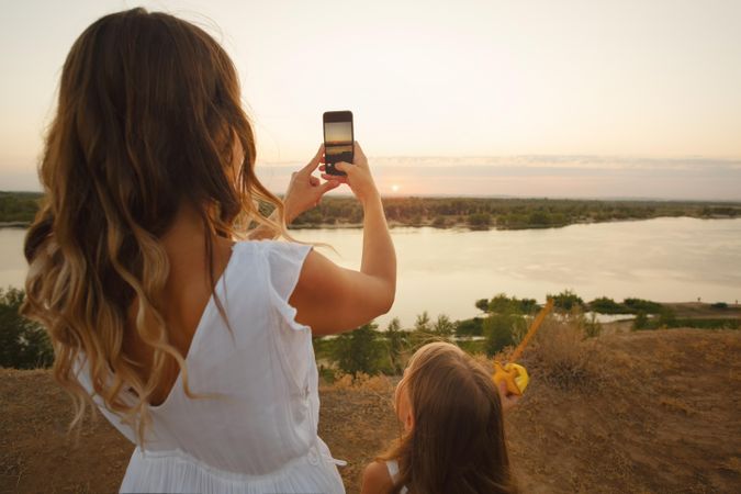 Woman taking photo of sunset with her phone on waterfront with little girl