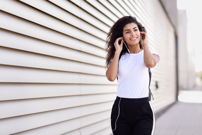 Arab woman in sport clothes with curly hair standing in front of wall adjusting headphones