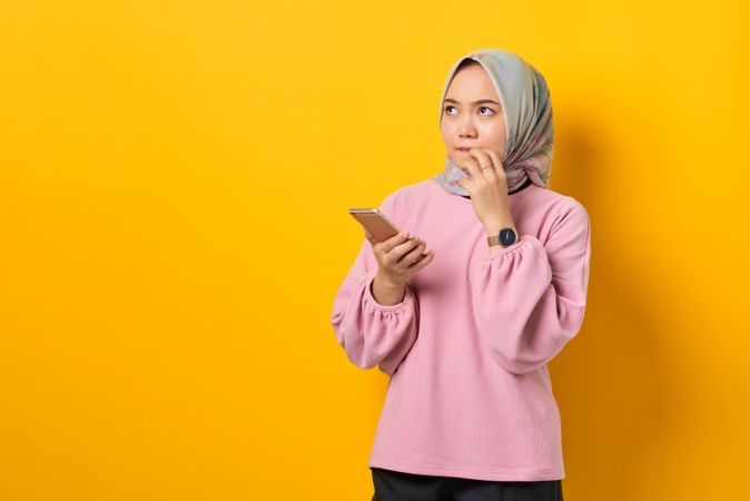 Muslim woman in headscarf holding smart phone and looking up in thought with fingers to mouth