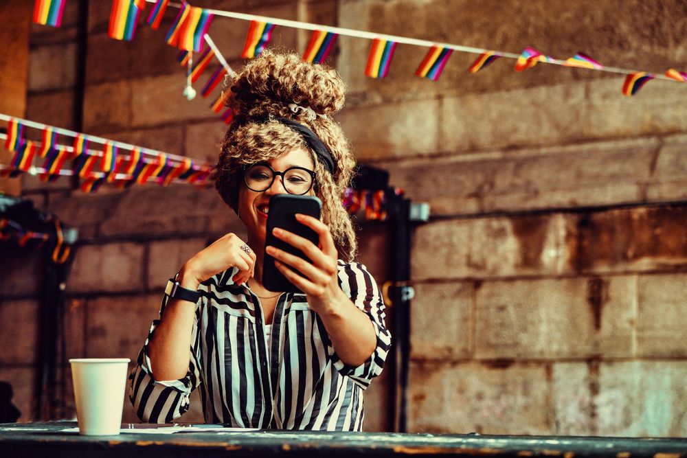 Laughing Black woman in stripe shirt taking selfie on phone in cafe