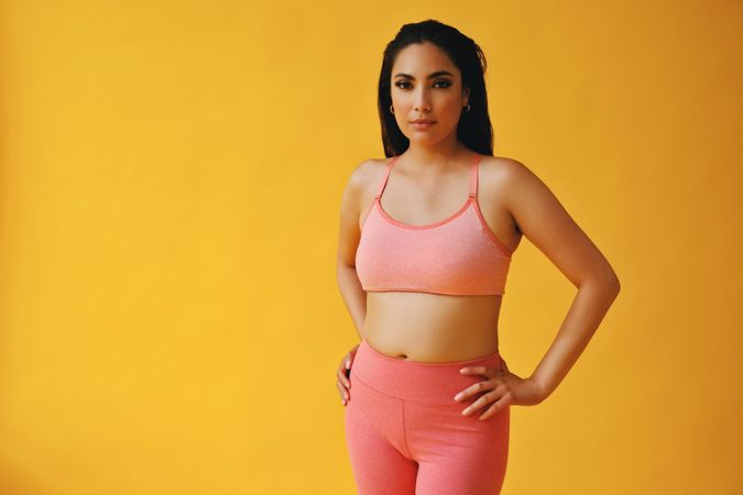 Hispanic female in yoga clothes standing in yellow studio with hands on hip, copy space