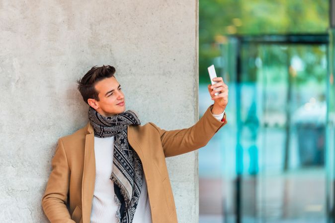 Stylish man leaning on wall outside taking selfie in camel coat with copy space