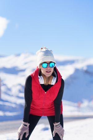 Front view of female in sunglasses and vest leaning on knees while recovering breath after run on winters day