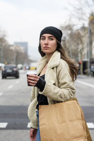 Portrait of young woman crossing the street, with takeaway coffee and shopping bag