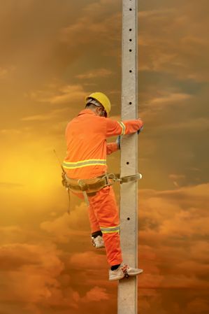 Back view of a worker in orange overall and yellow bump cap climbing a pole at sunset