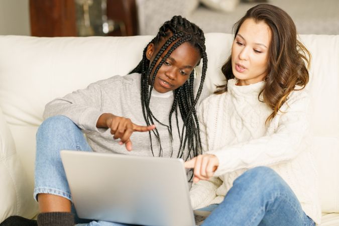 Two women in jeans and sweater pointing to something together on a sofa on a pc