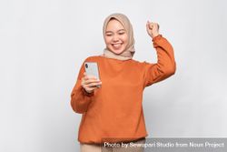 Muslim woman winning something in headscarf and orange shirt holding phone 56mrN0