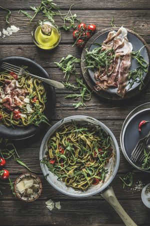 Italian food background with green cooked pasta in cooking pan with parma ham, arugula, tomatoes and cheese on rustic wooden kitchen table.