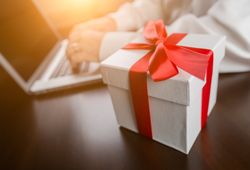 Gift Box with Red Ribbon and Bow Near Man Typing on Laptop Computer.
