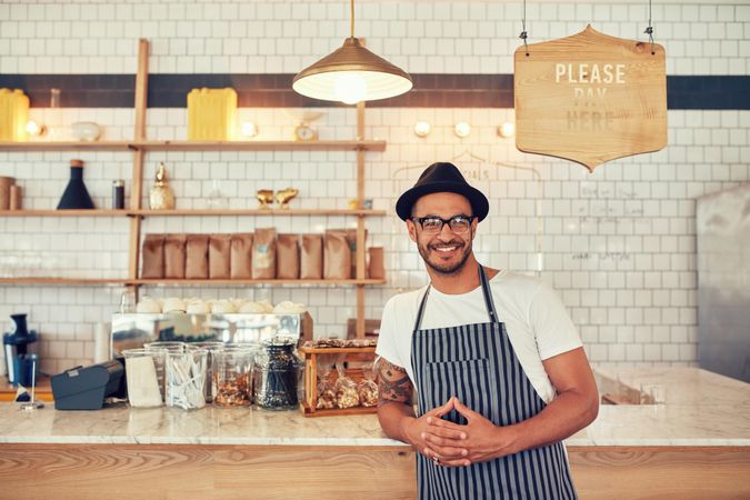 Portrait of happy young man wearing an apron and hat leaning to a cafe counter