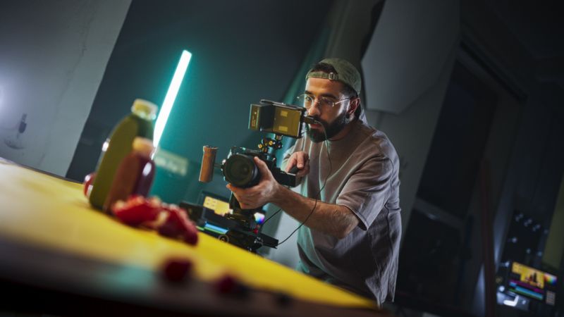 Focused Male Cinematographer With Beard, Wearing Cap And Glasses, Records Vibrant Scene On Professional Camera In Dimly Lit Studio, Capturing Essence Of Modern Videography And Storytelling.