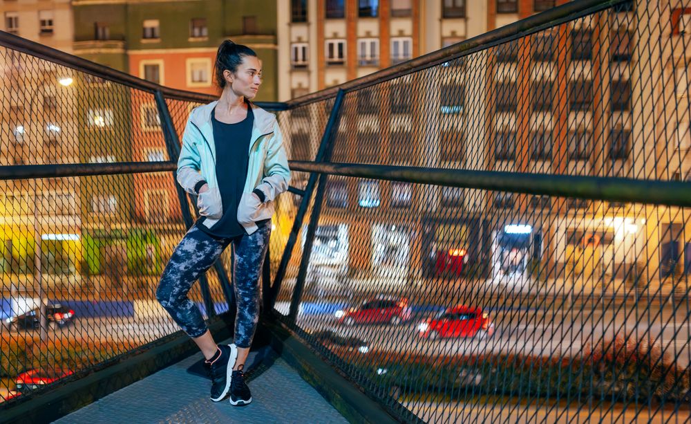 Pensive woman wearing casual sportswear standing on bridge illuminated by city lights at night