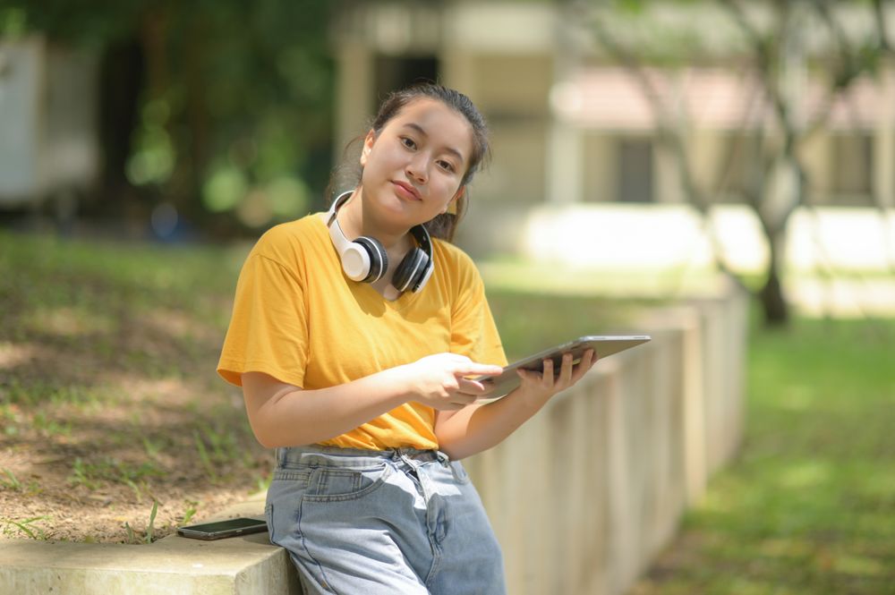 Woman in yellow shirt wearing headphones on neck holding tablet standing in garden looking at camera.