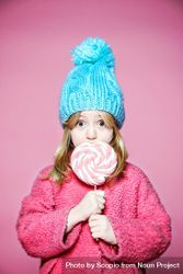 Girl in pink sweater and blue knit cap holding pink lollipop bxMAy0