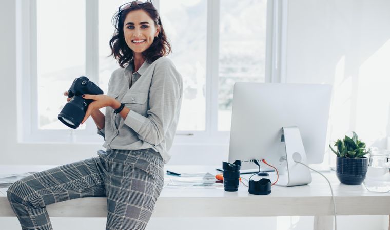 Freelance photographer sitting on office desk with digital camera in hand