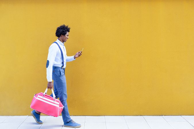 Side view of male with curly hair wearing shirt and suspenders walking next to yellow wall
