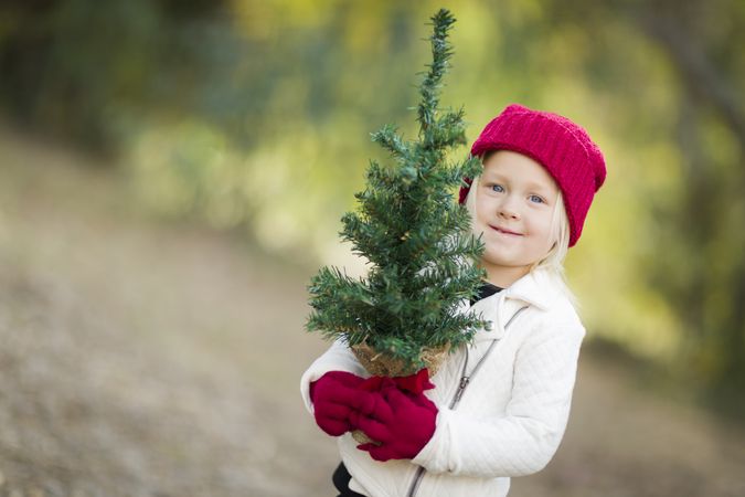Baby Girl In Red Mittens and Cap Holding Small Christmas Tree