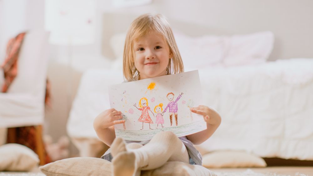 Young Smiling Girl Sitting on Pillows and Shows Drawing of Her Family.