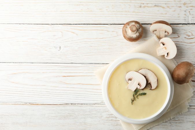 Looking down at bowl of creamy mushroom soup with herbs on napkin on wooden table, space for text