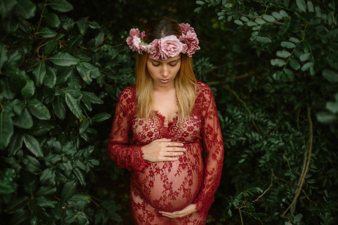 Pregnant woman with closed eyes touching belly and chin while standing in garden in sunny day