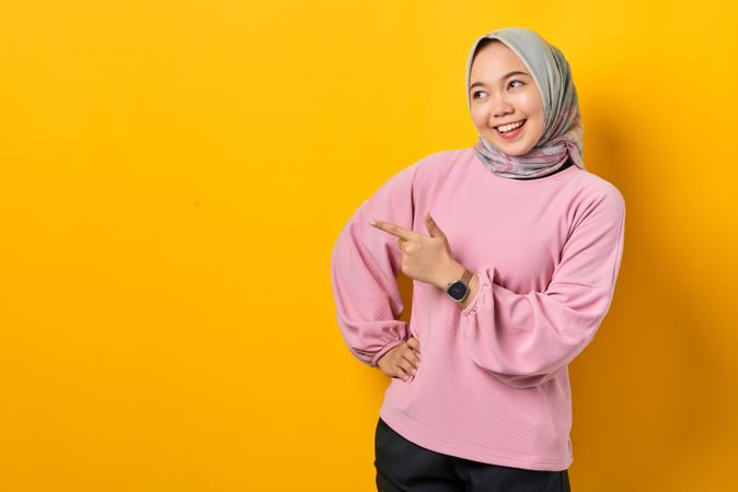 Smiling Muslim woman in scarf and pink sweater pointing towards copy space with hand on hip
