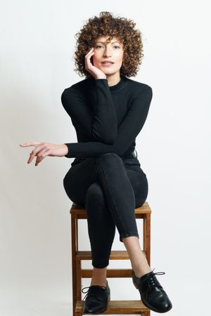 Woman dressed head to toe in dark color sitting on stool looking at camera resting head on hand