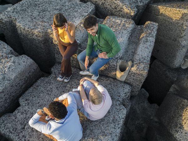 Aerial view of diverse friends sitting on rocks by the sea at sunset