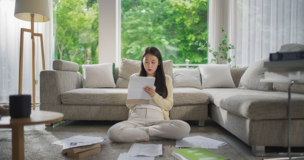 South Korean Woman Sitting on a Floor Next to a Cozy Couch, Working on University Homework