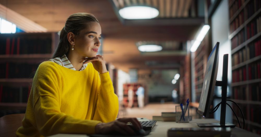 Female Student in Yellow Jumper Working on Her School Thesis on a Desktop Computer. Young Smart Woman Studying Online in a Quiet Public Library with Book Shelves