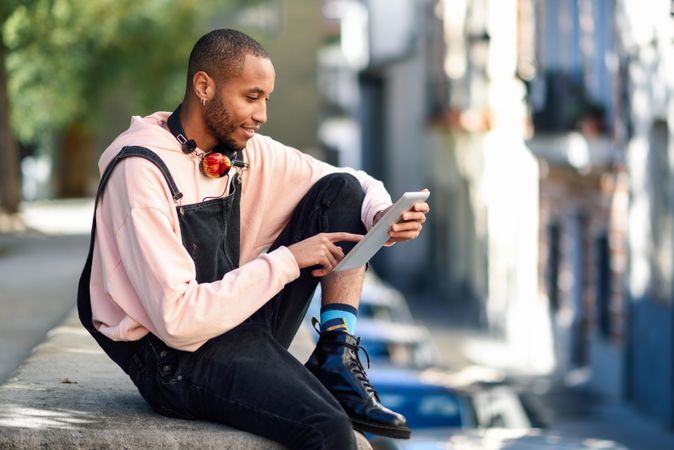 Calm male sitting with one leg up on wall in city checking tablet