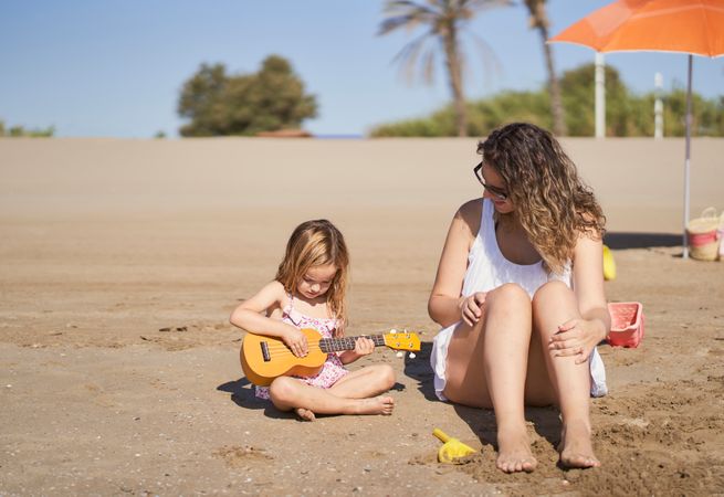 a little girl playing the ukulele sitting on the sand on the beach next to a mother