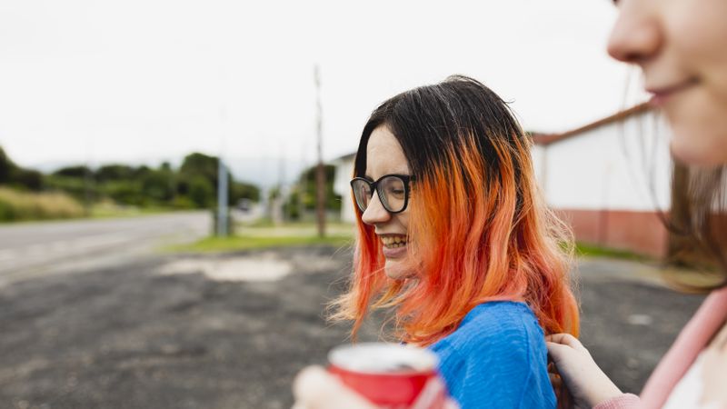 Two female friends, one with orange hair, chat and laugh while drinking a soda during a car stop
