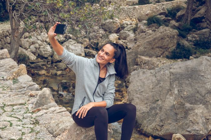 Cheerful young woman taking selfie while resting on stone