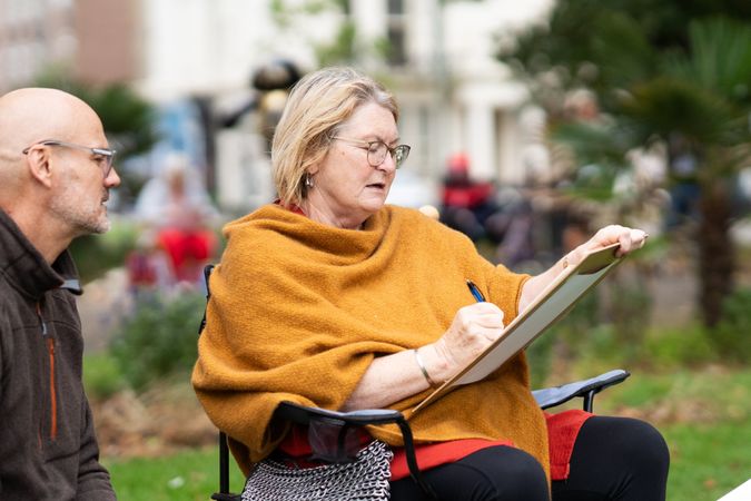 Woman sitting outside in park at drawing class with instructor over her shoulder