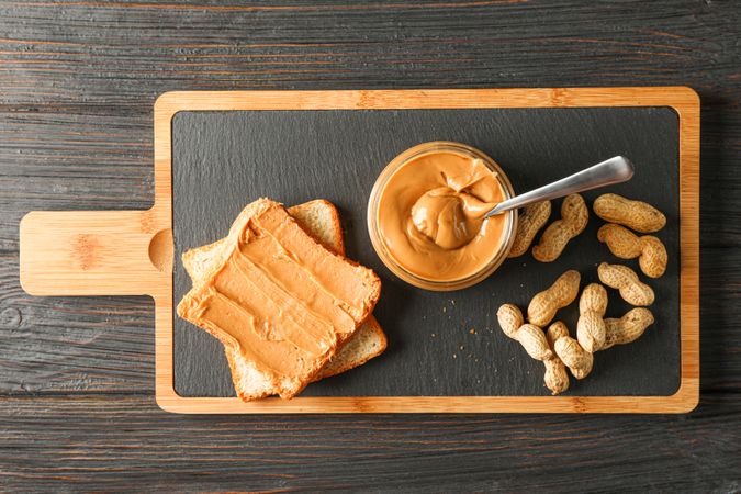 Glass jar with peanut butter and spoon, peanut, peanut butter sandwich and cutting board on wooden background, space for text and top view
