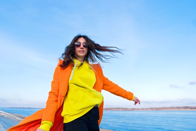Trendy woman in sunglasses, orange coat, yellow hoodie and jeans in front of the sea