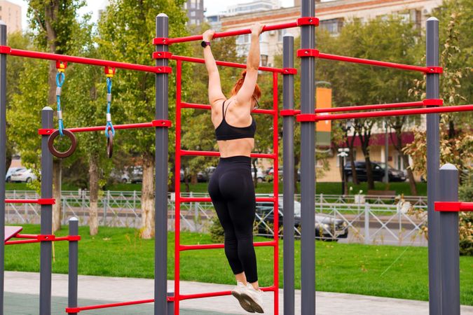 Woman performing pull-up on red outdoor fitness structure.