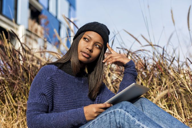 Questioning female in hat and sweater sitting outside with tablet