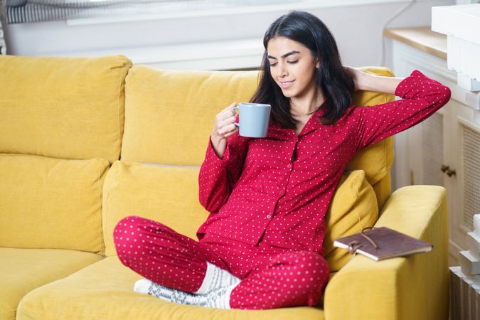 Female sipping tea on yellow sofa at home with notebook at her side