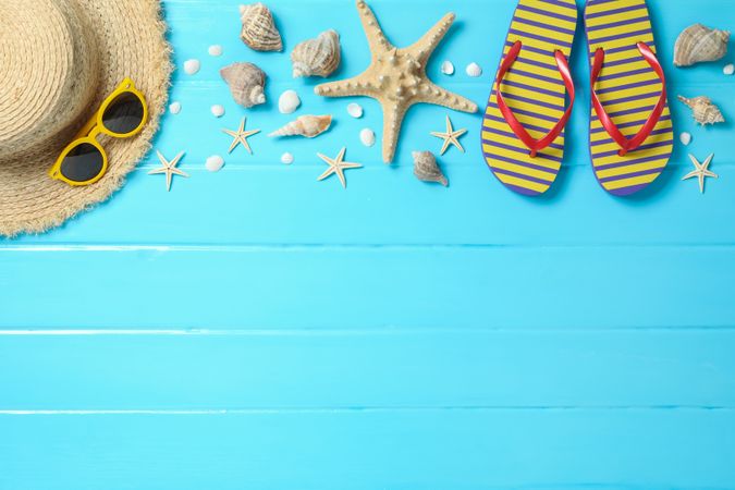 Straw hat, sunglasses, flip flops and many starfishes on color wooden background, space for text and top view. Summer vacation backdrop