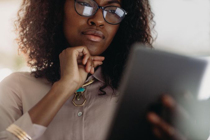 Close up of a curly haired businesswoman in spectacles looking at tablet pc with hand on chin