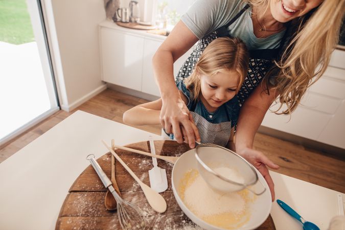 Cute girl sifting flour in bowl with her mother behind her