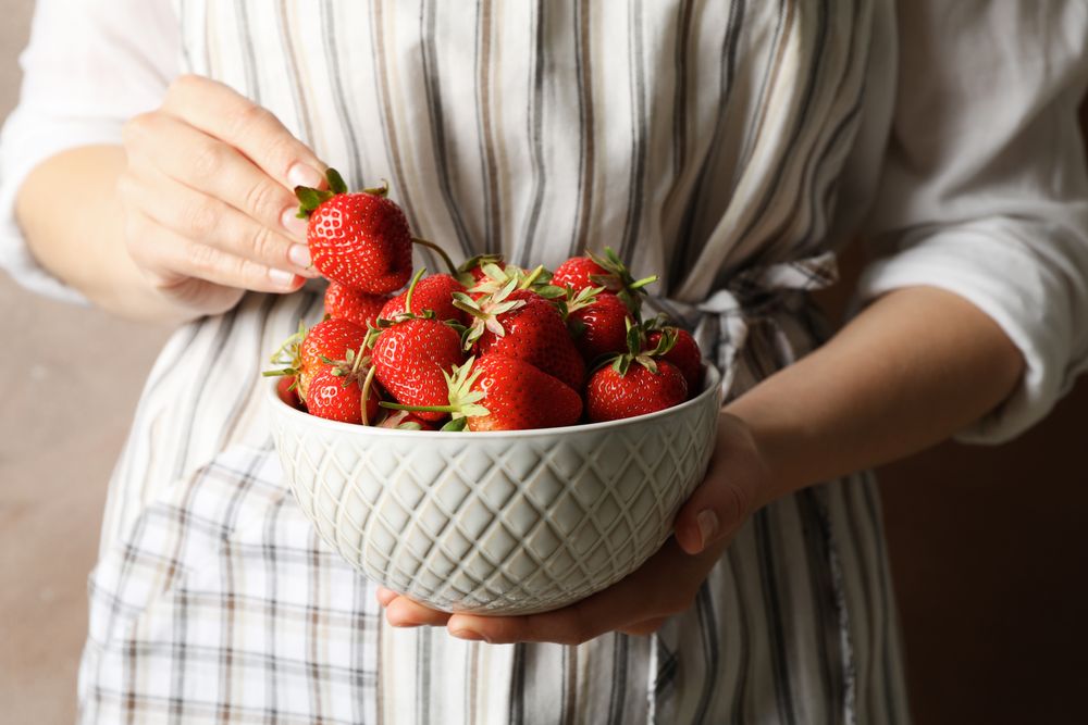 Woman holds bowl with tasty strawberry against brown background, close up.