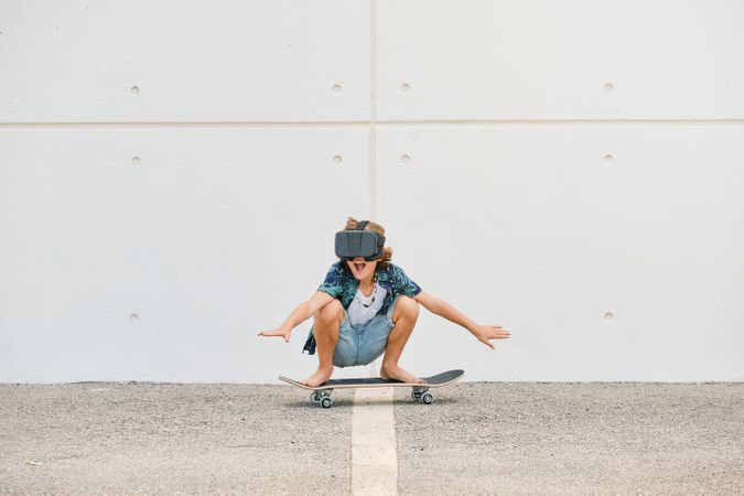 Cheerful child in VR glasses riding skateboard in city