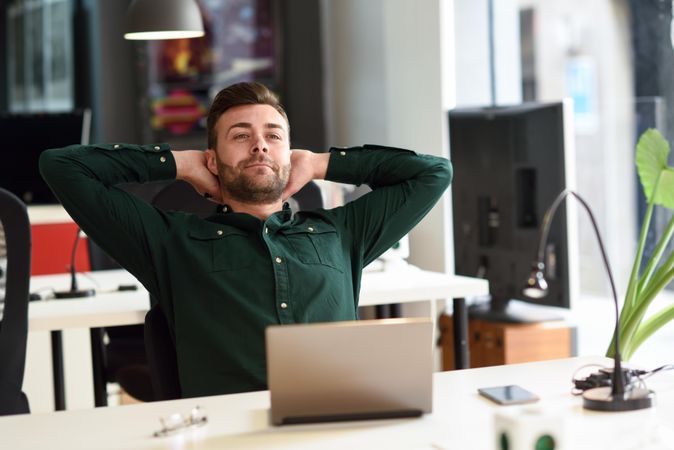 Young man with hands behind head at his computer in modern office