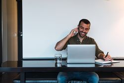 Cheerful man talking on smartphone near laptop