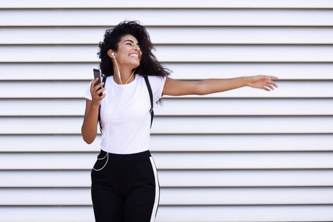 Arab woman in sport clothes with curly hair standing in front of wall dancing with phone