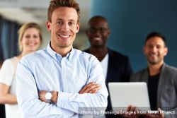 Smiling man with arms crossed pictures in front of colleagues 0L2GD4